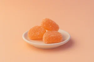 Three sugar-coated orange slice candies arranged on a small white ceramic dish, set against a soft pastel peach background, evoking nostalgia, warmth, and childhood comfort.